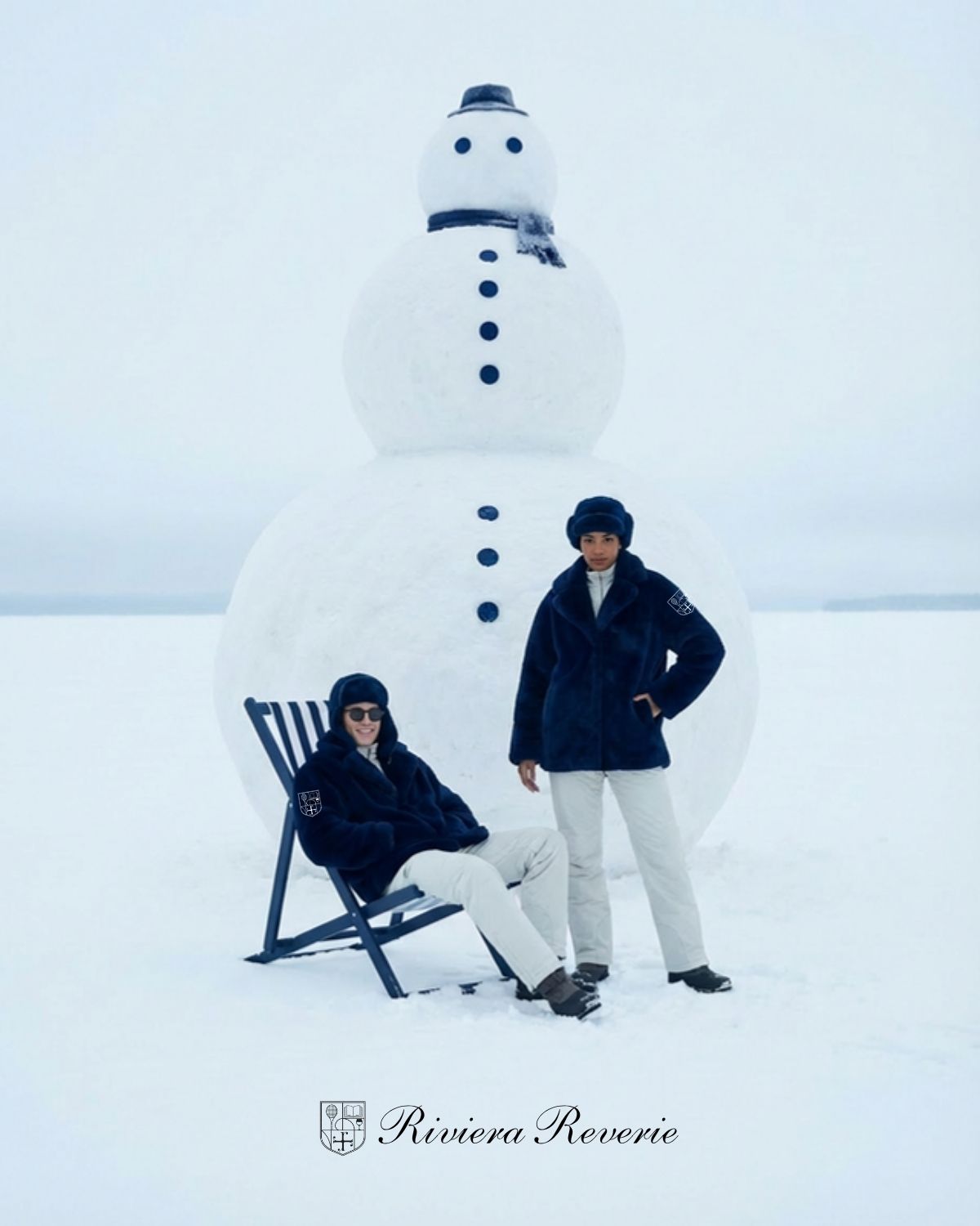 Two people in navy coats and Riviera Reverie hats beside a giant snowman on a snowfield